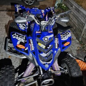 May include: A blue Yamaha ATV with black and white camouflage graphics. The ATV has a GoPro camera mounted on the front and a set of aftermarket handlebars. The ATV is sitting on a paved surface.