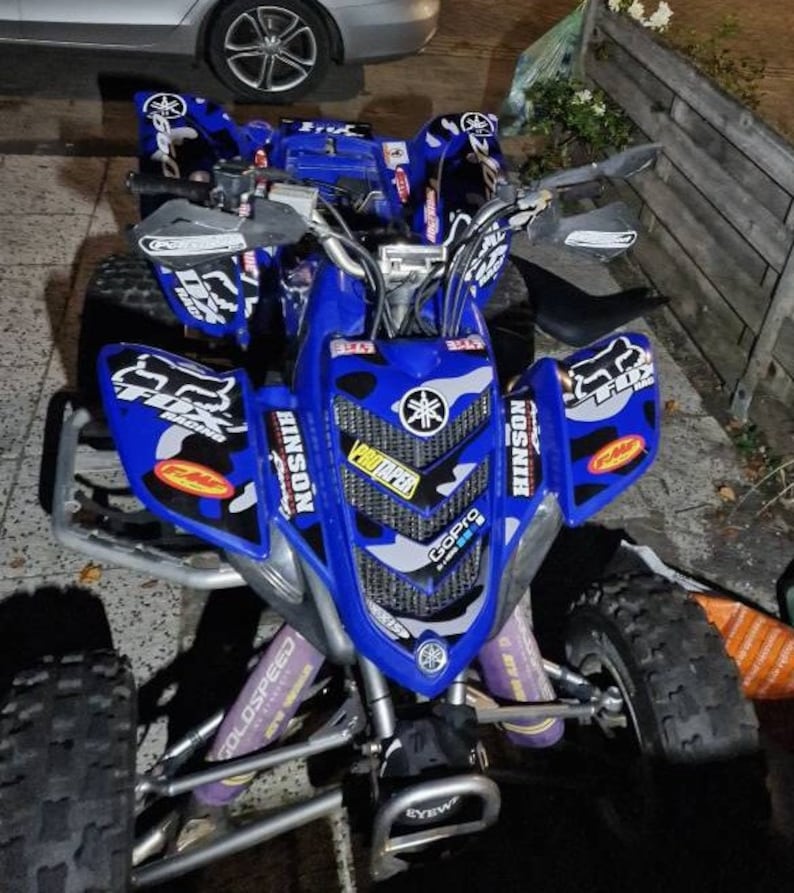 May include: A blue Yamaha ATV with black and white camouflage graphics. The ATV has a GoPro camera mounted on the front and a set of aftermarket handlebars. The ATV is sitting on a paved surface.