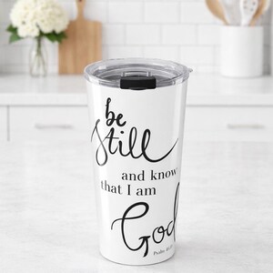 May include: White travel tumbler with a clear lid and black text that reads "be still and know that I am God" from Psalm 46:10. The tumbler is cylindrical and appears to be made of stainless steel.