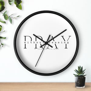 Op de afbeelding: Een ronde wandklok met een zwart frame en een witte wijzerplaat. De klok heeft zwarte wijzers en de woorden "PRAY without ceasing" in een vetgedrukte, zwarte lettertype. De tekst "1 Thessalonians" staat eronder.