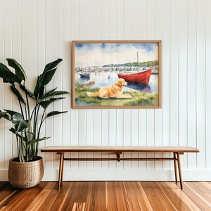 Puede incluir: Una pintura de acuarela de un perro golden retriever sentado en una orilla cubierta de hierba, mirando un bote rojo en un puerto. La pintura est&aacute; enmarcada en un marco de madera claro y cuelga de una pared blanca.