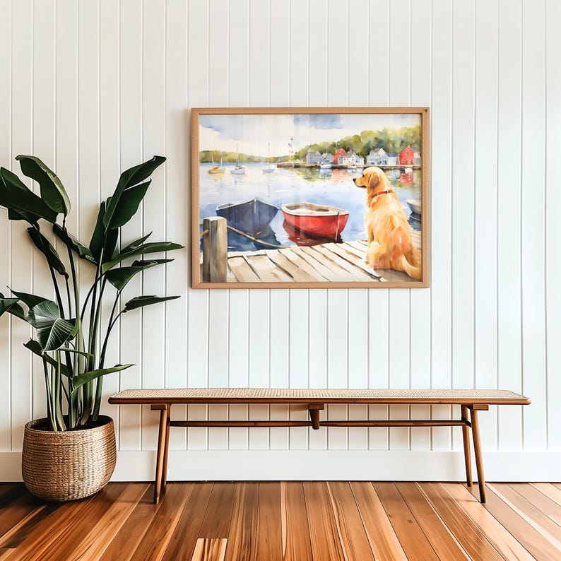 Puede incluir: Una pintura al agua enmarcada de un perro golden retriever sentado en un muelle de madera con vistas a un peque&ntilde;o puerto con barcos. La pintura est&aacute; colgada en una pared blanca con un banco de madera en primer plano.