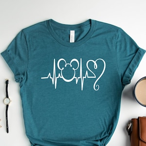 Op de afbeelding: Tealblauw t-shirt met een witte afbeelding van een Mickey Mouse-hoofd met een hartslaglijn en een hart. Het shirt ligt plat op een wit oppervlak met een horloge, bril, oorbellen, een kopje koffie en een bruine handtas.