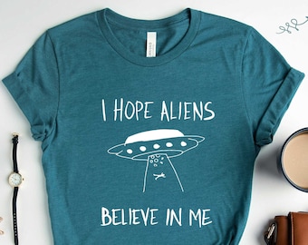 Puede incluir: Una camiseta de color verde azulado jaspeado con el texto "Espero que los extraterrestres crean en mí" y un dibujo de un OVNI.