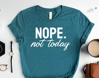 以下が含まれることがあります： 白地に「NOPE. not today」という白い文字が書かれた、ターコイズ色のヘザー素材のTシャツ。Tシャツは、コーヒーカップ、茶色の革製のバッグ、腕時計、眼鏡、そして小さな黒いトレイにイヤリングが置かれた白い表面の上に平らに置かれている。