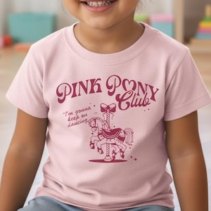 May include: A light pink t-shirt featuring the text "Pink Pony Club" in a burgundy script, along with a carousel horse graphic. The shirt is worn by a young child with dark hair styled in two buns.