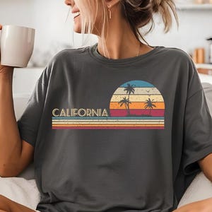 Puede incluir: Una mujer lleva una camiseta gris con un gr&aacute;fico de puesta de sol vintage y el texto "CALIFORNIA" en blanco. El gr&aacute;fico presenta palmeras y una puesta de sol con colores naranja, amarillo, rojo y azul.