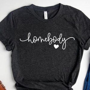 May include: A dark gray t-shirt with the text "homebody" in white cursive lettering and a small white heart. The shirt is laid flat on a white surface with a pair of jeans, a small brown purse, a pair of glasses, and a camera.