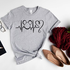 Op de afbeelding: Een grijs T-shirt met een zwart Mickey Mouse-hoofd en een hartslagontwerp. Het ontwerp bevat een zwart hart met een krul aan de bovenkant.