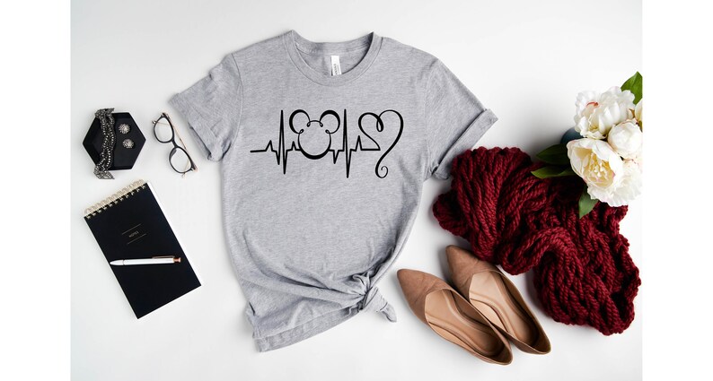 Op de afbeelding: Een grijs T-shirt met een zwart Mickey Mouse-hoofd en een hartslagontwerp. Het ontwerp bevat een zwart hart met een krul aan de bovenkant.