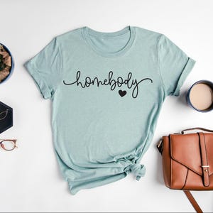 May include: A light blue t-shirt with the text "homebody" in black cursive lettering and a small black heart.