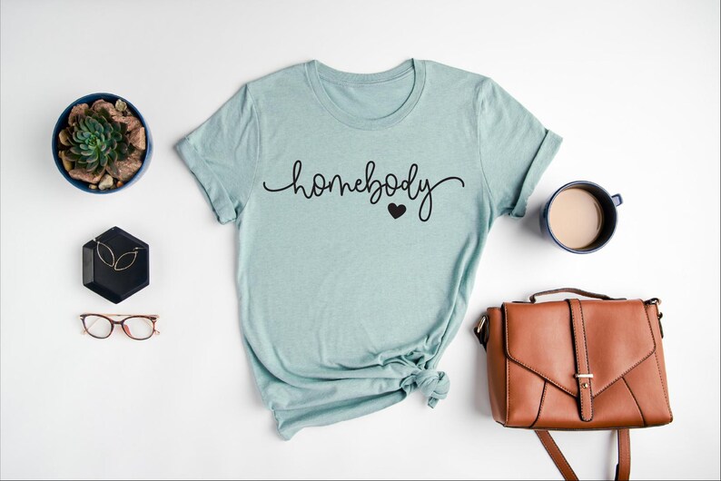 May include: A light blue t-shirt with the text "homebody" in black cursive lettering and a small black heart.