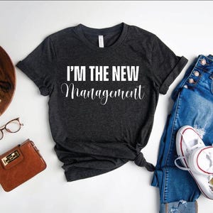 Pu&ograve; includere: Una maglietta grigia scura con la scritta "I'm the new management" stampata in bianco sul davanti. La maglietta &egrave; stesa su una superficie bianca con un paio di jeans, una borsa e una macchina fotografica.