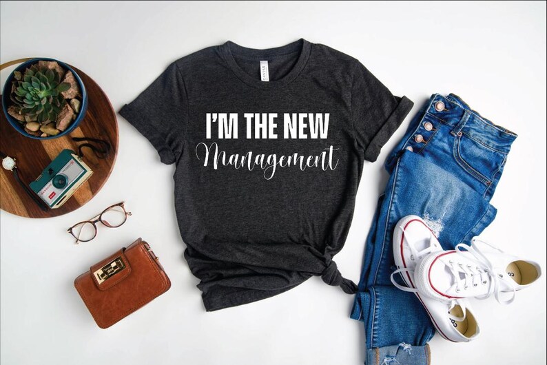 Pu&ograve; includere: Una maglietta grigia scura con la scritta "I'm the new management" stampata in bianco sul davanti. La maglietta &egrave; stesa su una superficie bianca con un paio di jeans, una borsa e una macchina fotografica.