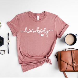 May include: A dusty rose t-shirt with the text "homebody" in white script with a small white heart.