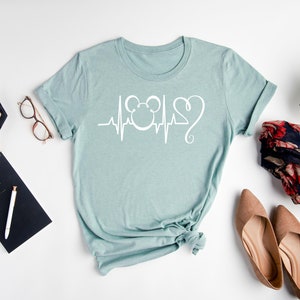 Op de afbeelding: Een lichtblauw t-shirt met een witte afbeelding van een Mickey Mouse-hoofd en een hartslaglijn. De afbeelding bevat ook een hart met een swirl.