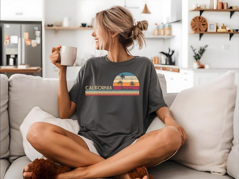 Puede incluir: Una mujer lleva una camiseta gris con un gr&aacute;fico de puesta de sol vintage y el texto "CALIFORNIA" en blanco. El gr&aacute;fico presenta palmeras y una puesta de sol con colores naranja, amarillo, rojo y azul.
