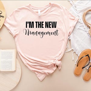 Pu&ograve; includere: Una maglietta rosa con il testo "I'm the new management" in lettere nere.