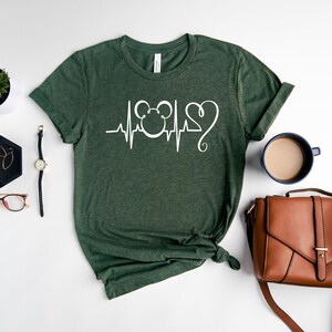 Op de afbeelding: Een groen T-shirt met een witte afbeelding van een hartslag met een Mickey Mouse-hoofd en een hart.