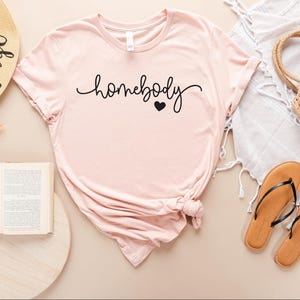 May include: A pink t-shirt with the text "homebody" in black cursive lettering and a small black heart.