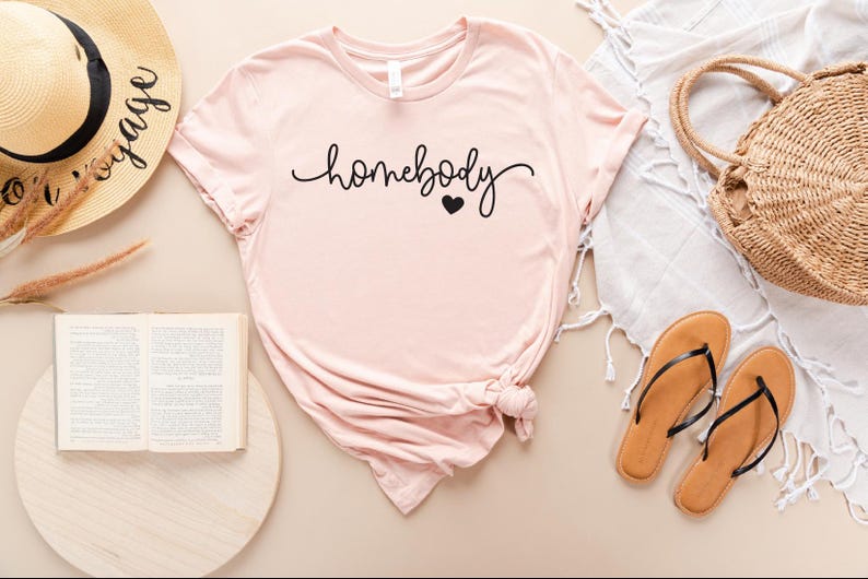 May include: A pink t-shirt with the text "homebody" in black cursive lettering and a small black heart.