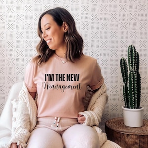 Pu&ograve; includere: Una donna indossa una maglietta rosa con il testo "I'm the new management" in lettere nere. &Egrave; seduta su una sedia con un cardigan bianco e pantaloni bianchi.