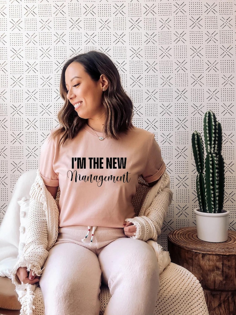 Pu&ograve; includere: Una donna indossa una maglietta rosa con il testo "I'm the new management" in lettere nere. &Egrave; seduta su una sedia con un cardigan bianco e pantaloni bianchi.
