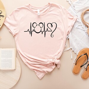 Op de afbeelding: Een roze T-shirt met een zwarte afbeelding van een Mickey Mouse-hoofd met een hartslag en een hart. Het shirt ligt plat op een lichtbruin oppervlak.