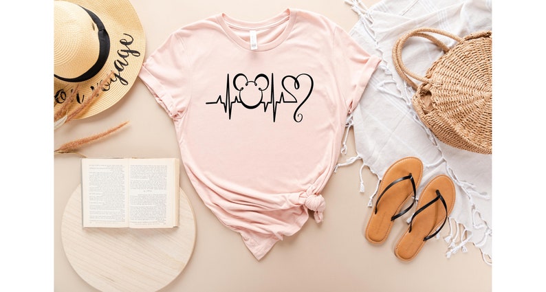 Op de afbeelding: Een roze T-shirt met een zwarte afbeelding van een Mickey Mouse-hoofd met een hartslag en een hart. Het shirt ligt plat op een lichtbruin oppervlak.