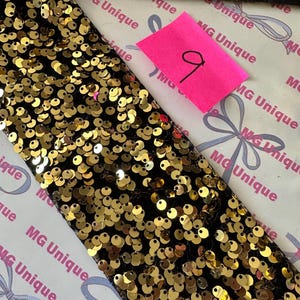 Op de afbeelding: Detailopname van een zwarte stoffen strook, dicht bedekt met gouden pailletten. Een roze plakbriefje met het nummer "9" in zwarte inkt is zichtbaar. De achtergrond toont zwarte stof en papier met "MG Unique".