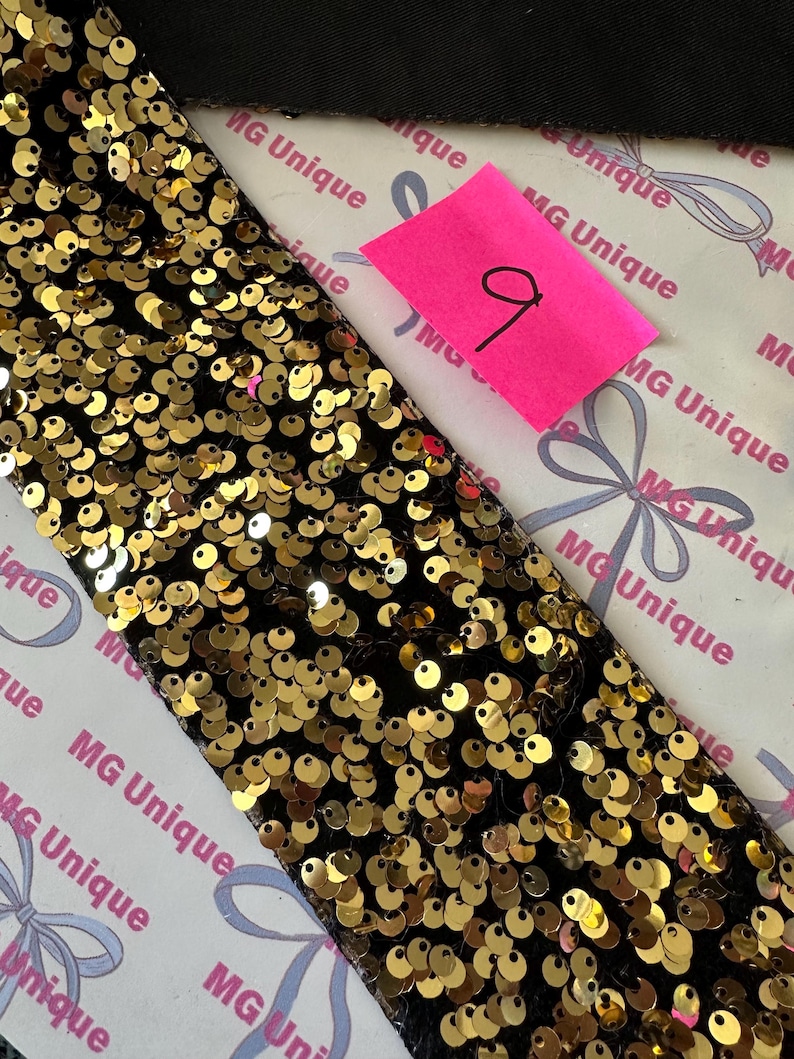 Op de afbeelding: Detailopname van een zwarte stoffen strook, dicht bedekt met gouden pailletten. Een roze plakbriefje met het nummer "9" in zwarte inkt is zichtbaar. De achtergrond toont zwarte stof en papier met "MG Unique".