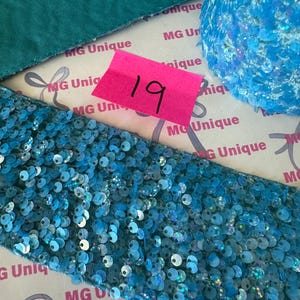 Op de afbeelding: Close-up van verschillende blauwe paillettenlinten en stof. Een roze label met het nummer 19 is zichtbaar. De linten hebben verschillende vormen en texturen, met een turquoise stof op de achtergrond. De linten zijn versierd met glanzende, iriserende pailletten.