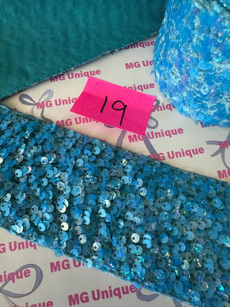 Op de afbeelding: Close-up van verschillende blauwe paillettenlinten en stof. Een roze label met het nummer 19 is zichtbaar. De linten hebben verschillende vormen en texturen, met een turquoise stof op de achtergrond. De linten zijn versierd met glanzende, iriserende pailletten.