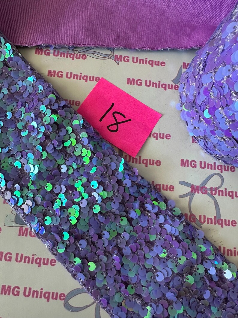 Op de afbeelding: Close-up van paarse paillettenstof met iriserende groene en blauwe accenten. Een roze label met het nummer 18 is zichtbaar. De stof is waarschijnlijk voor knutselen of versiering.