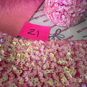 Op de afbeelding: Roze paillettenlint en een rol roze paillettenstof. Aan de linkerkant is een roze lint zichtbaar. In het midden zit een roze label met het nummer 21. De paillettenstof is lichtroze.