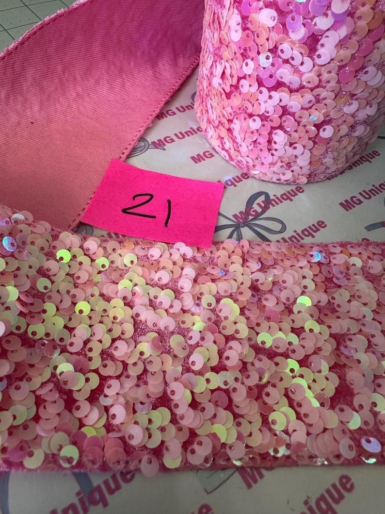Op de afbeelding: Roze paillettenlint en een rol roze paillettenstof. Aan de linkerkant is een roze lint zichtbaar. In het midden zit een roze label met het nummer 21. De paillettenstof is lichtroze.