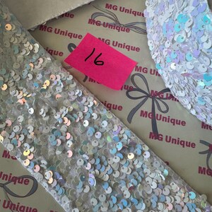 Op de afbeelding: Close-up van witte stof bedekt met pailletten en een bijpassend accessoire. De pailletten hebben een iriserende glans, die verschillende kleuren reflecteert. Een roze label met het nummer 16 is zichtbaar. De achtergrond toont de tekst "MG Unique".