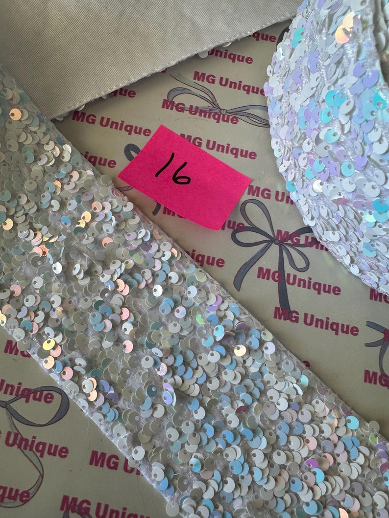 Op de afbeelding: Close-up van witte stof bedekt met pailletten en een bijpassend accessoire. De pailletten hebben een iriserende glans, die verschillende kleuren reflecteert. Een roze label met het nummer 16 is zichtbaar. De achtergrond toont de tekst "MG Unique".