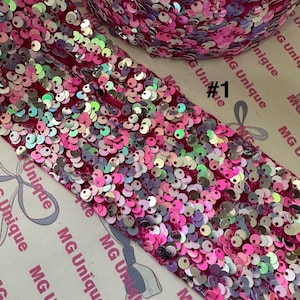 Op de afbeelding: Close-up van een rol roze paillettenstof met iriserende pailletten. De stof heeft een dieproze kleur en de pailletten zijn een mix van roze, zilver en iriserende kleuren. De tekst "#1" is zichtbaar.