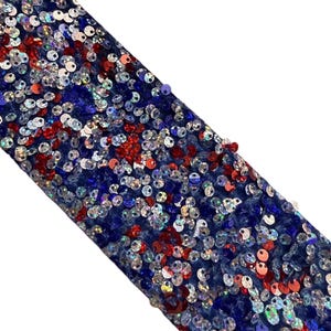 Op de afbeelding: Close-up van een stoffen strook versierd met rode, witte en blauwe pailletten. De pailletten zijn dicht op een donkerblauwe stof geplaatst, wat een patriottisch patroon cre&euml;ert. Diagonale weergave.