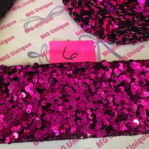 Op de afbeelding: Close-up van een strook fuchsia pailletten op een zwarte achtergrond. De pailletten zijn dicht op elkaar gepakt, waardoor een glinsterend effect ontstaat. Een roze label met het nummer 6 is zichtbaar. De achtergrond heeft een herhalend patroon met de tekst "MG Unique".
