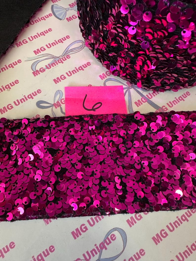Op de afbeelding: Close-up van een strook fuchsia pailletten op een zwarte achtergrond. De pailletten zijn dicht op elkaar gepakt, waardoor een glinsterend effect ontstaat. Een roze label met het nummer 6 is zichtbaar. De achtergrond heeft een herhalend patroon met de tekst "MG Unique".