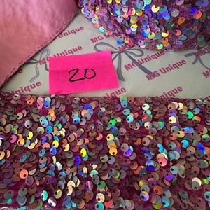 Op de afbeelding: Close-up van een rol iriserende paillettenbies in roze en paarse tinten. Een roze label met het nummer "20" is zichtbaar. De bies ligt op een oppervlak met de tekst "MG Unique" erop.