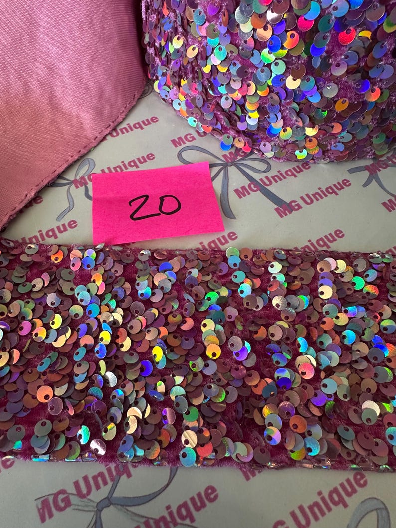 Op de afbeelding: Close-up van een rol iriserende paillettenbies in roze en paarse tinten. Een roze label met het nummer "20" is zichtbaar. De bies ligt op een oppervlak met de tekst "MG Unique" erop.