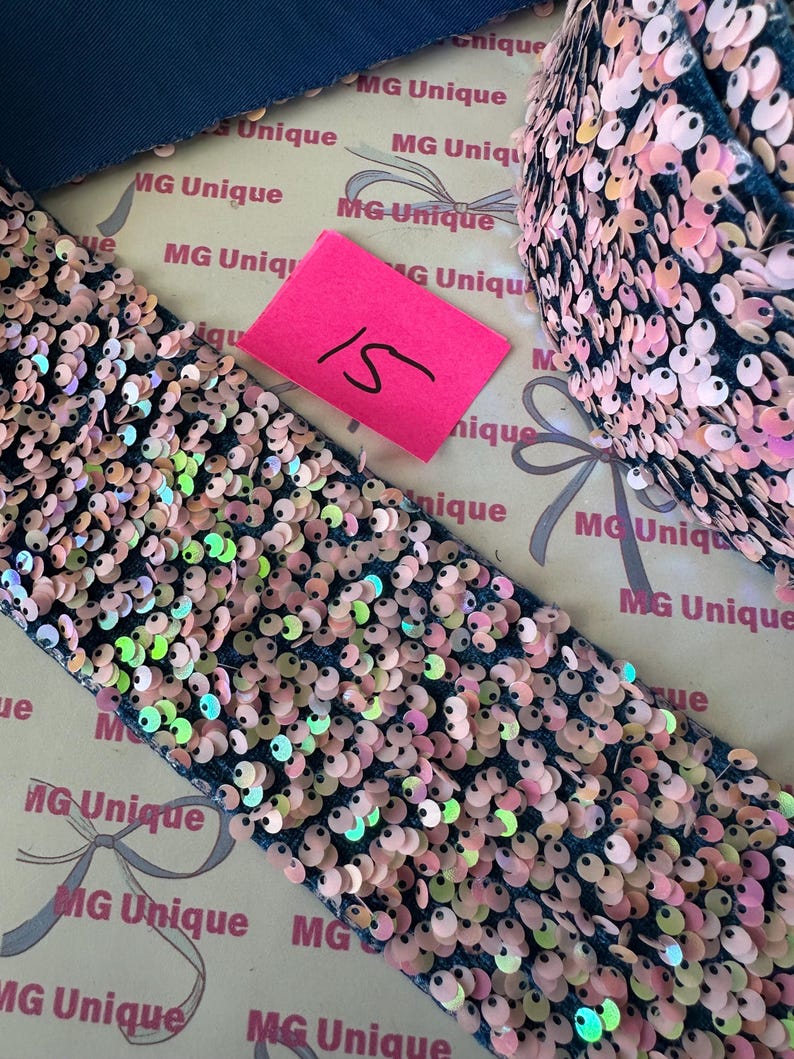 Op de afbeelding: Close-up van een donkerblauwe stof versierd met iriserende roze pailletten. Een roze label met het nummer 15 is zichtbaar. Geschikt voor knutselen of decoratie.