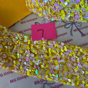 Op de afbeelding: Close-up van gele paillettenlint met iriserende accenten. Een effen geel lint is zichtbaar naast het paillettenlint. Een roze label met het nummer 7 staat in het midden van de afbeelding. De achtergrond heeft de tekst "MG Unique".