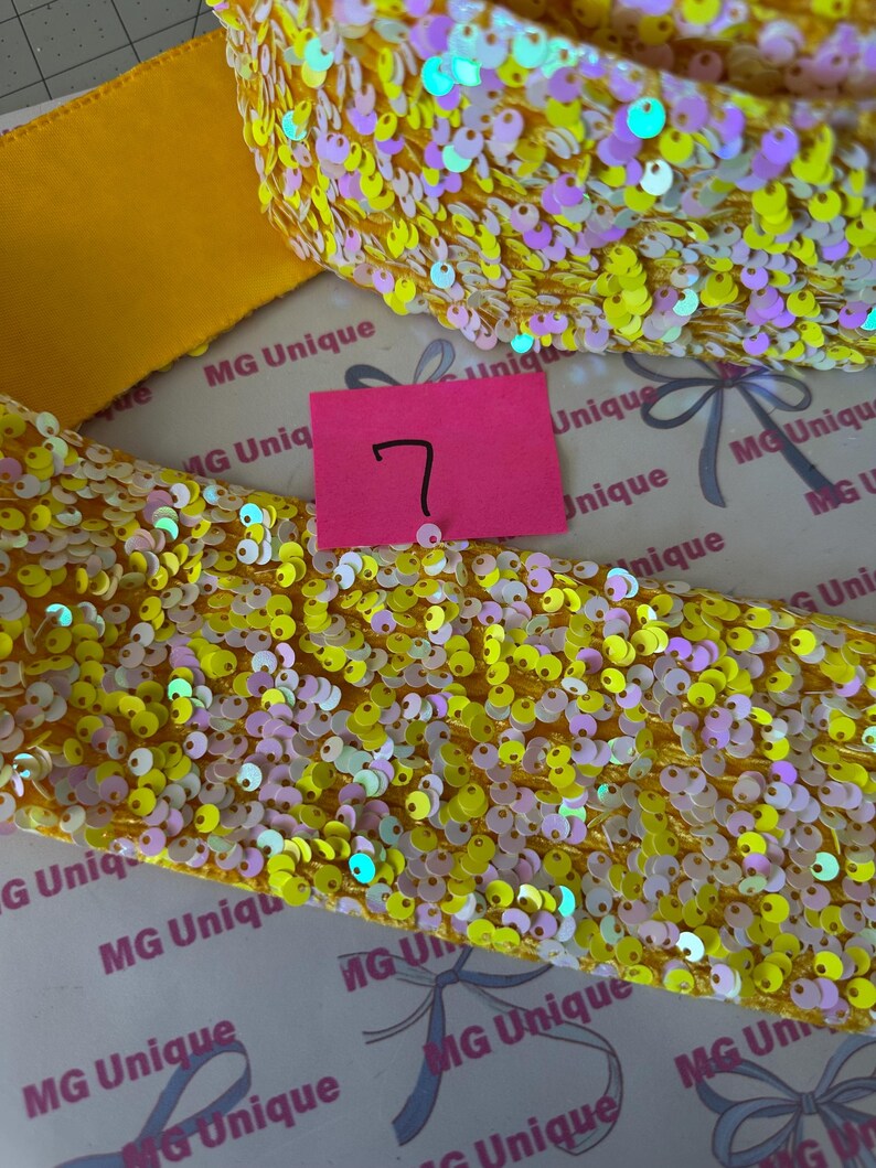 Op de afbeelding: Close-up van gele paillettenlint met iriserende accenten. Een effen geel lint is zichtbaar naast het paillettenlint. Een roze label met het nummer 7 staat in het midden van de afbeelding. De achtergrond heeft de tekst "MG Unique".