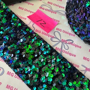 Op de afbeelding: Close-up van een rol zwarte paillettenbies met groen en blauw iriserende pailletten. Een roze label met het nummer 12 is zichtbaar. De bies ligt op een witte achtergrond met de tekst "MG Unique" herhaaldelijk afgedrukt.