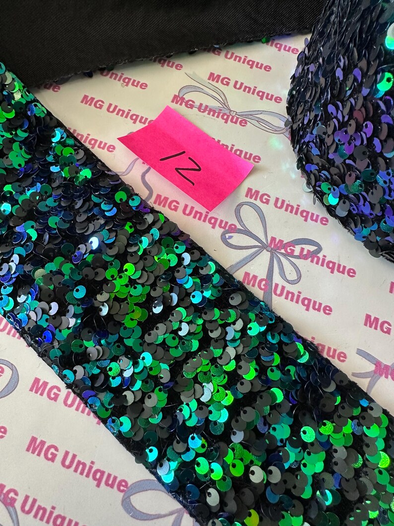 Op de afbeelding: Close-up van een rol zwarte paillettenbies met groen en blauw iriserende pailletten. Een roze label met het nummer 12 is zichtbaar. De bies ligt op een witte achtergrond met de tekst "MG Unique" herhaaldelijk afgedrukt.