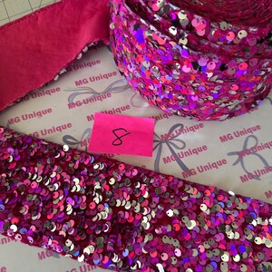Op de afbeelding: Een rol levendig roze paillettenlint en een stuk paillettenlint worden getoond. Het lint is bedekt met glanzende, iriserende pailletten. Een kleiner stuk effen roze lint is ook zichtbaar. Een roze plakbriefje met het nummer 8 staat in de afbeelding.